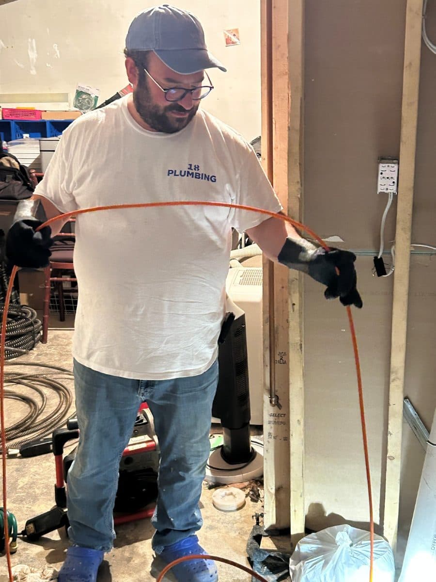 Plumber in an 18 Plumbing shirt running a drain machine into a basement floor drain.